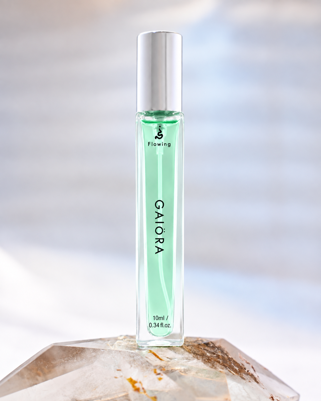 A 10ml clear glass roller bottle with a silver cap, filled with a light mint green liquid, standing on a faceted crystal against a soft, blurred light blue and white background. The brand name 'Flowing' and scent 'GAIORA' are visible on the bottle.