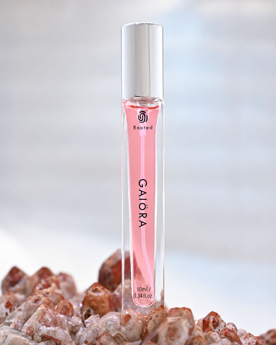 A clear cylindrical perfume or roller bottle filled with pink liquid, standing upright on a bed of rough pink and white crystal. The bottle has a silver cap and visible branding reading 'Rooted' and 'GAIORA'.